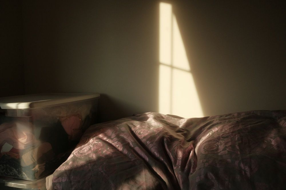 A dim bedroom at dawn. A single shaft of sun falls on the wall above an unmade bed.