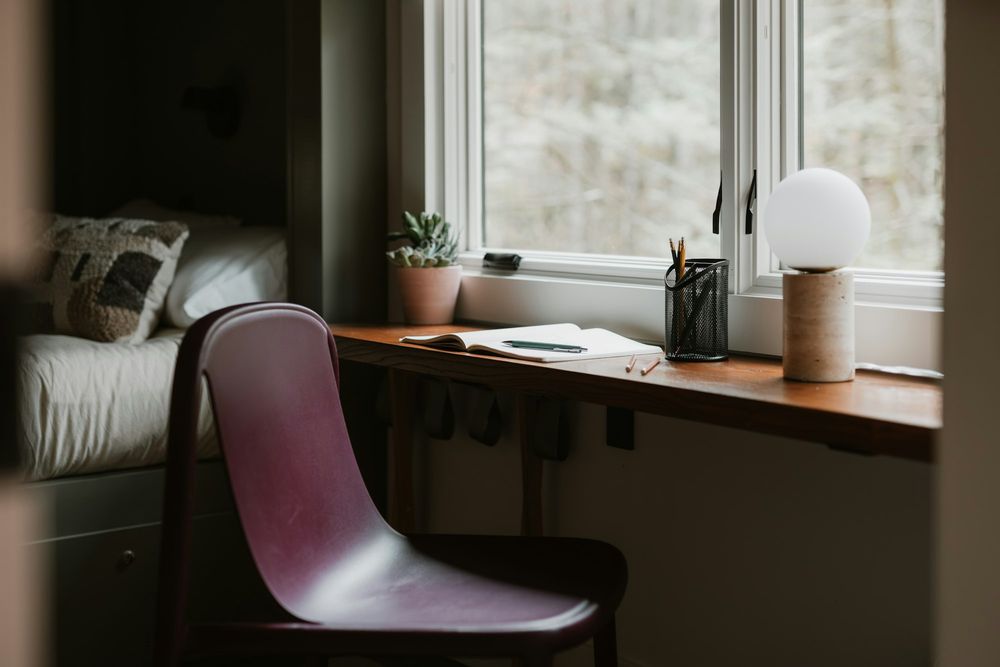 A narrow desk along a window, with an open notebook, a pencil pot and a small globe lamp; a chair pulled up to it.