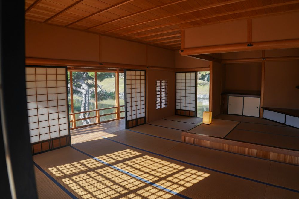 A wide tatami room with shoji screens open to a garden; afternoon sun draws a lattice of light on the mats.