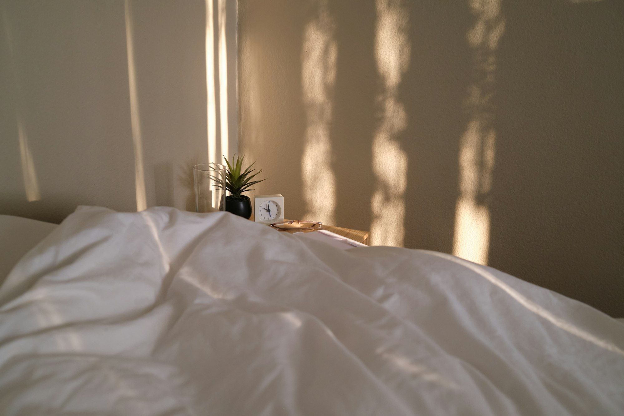 A white duvet in early sun, with long shadows of leaves falling on the bedroom wall.