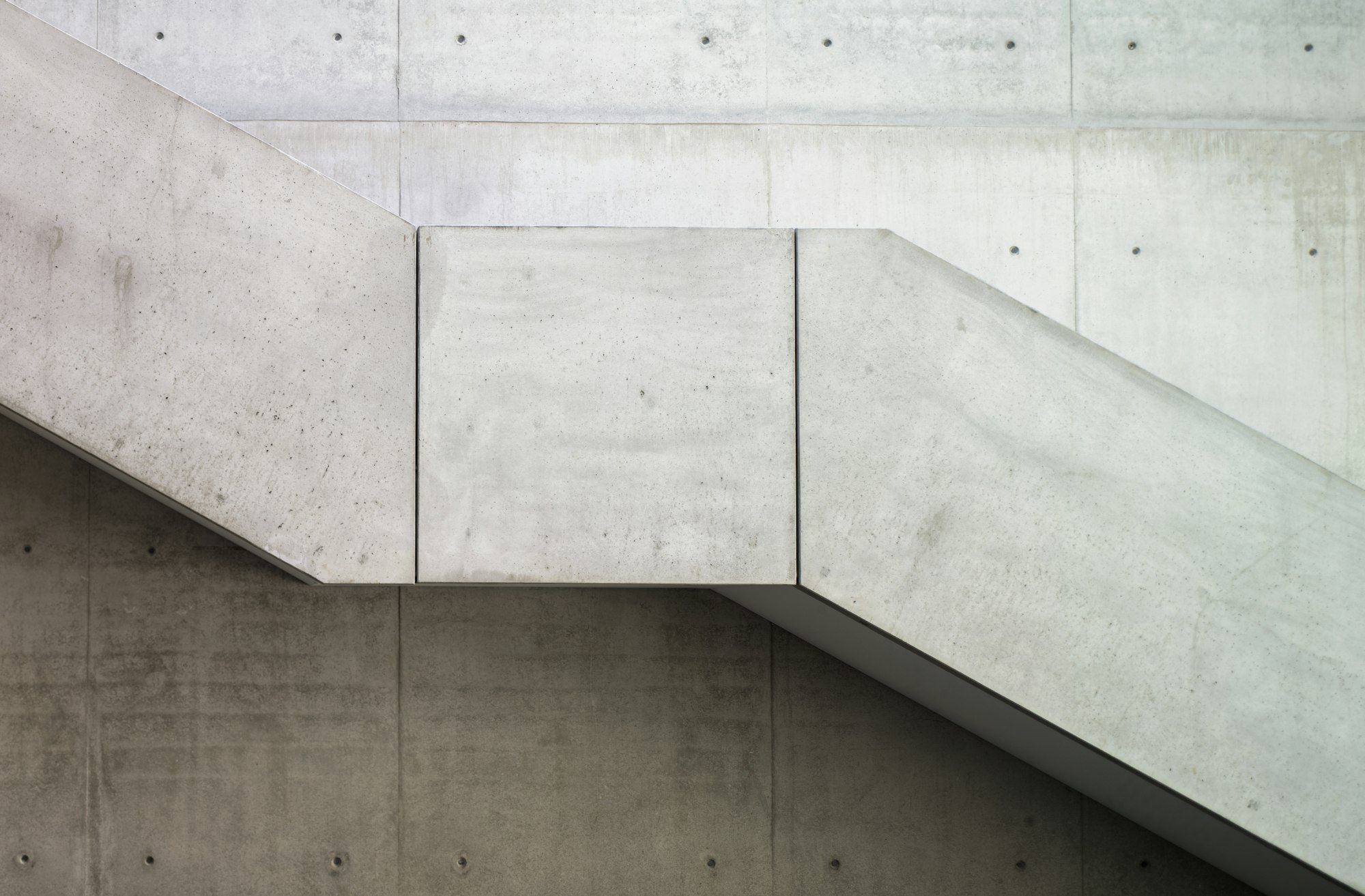 A fair-faced concrete stair climbing across a concrete wall with rows of tie holes, in soft daylight.