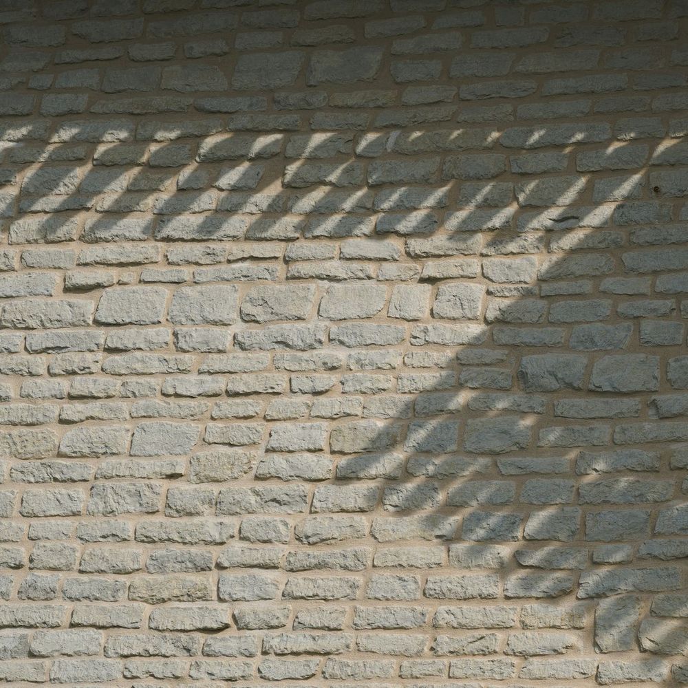 A pale brick wall under grazing light, striped by the shadow of a grille.