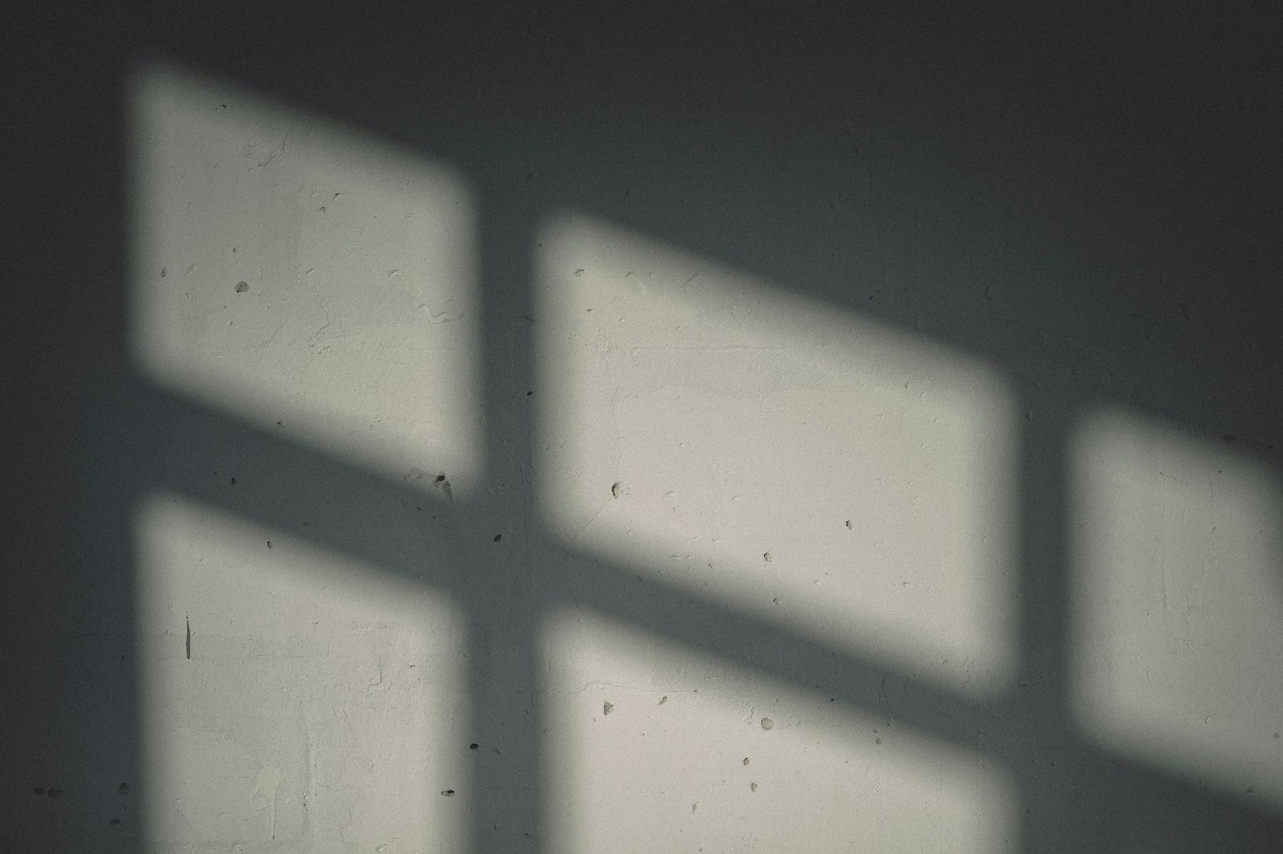The shadow of a window frame falls as a cross of light on a pale plaster wall.
