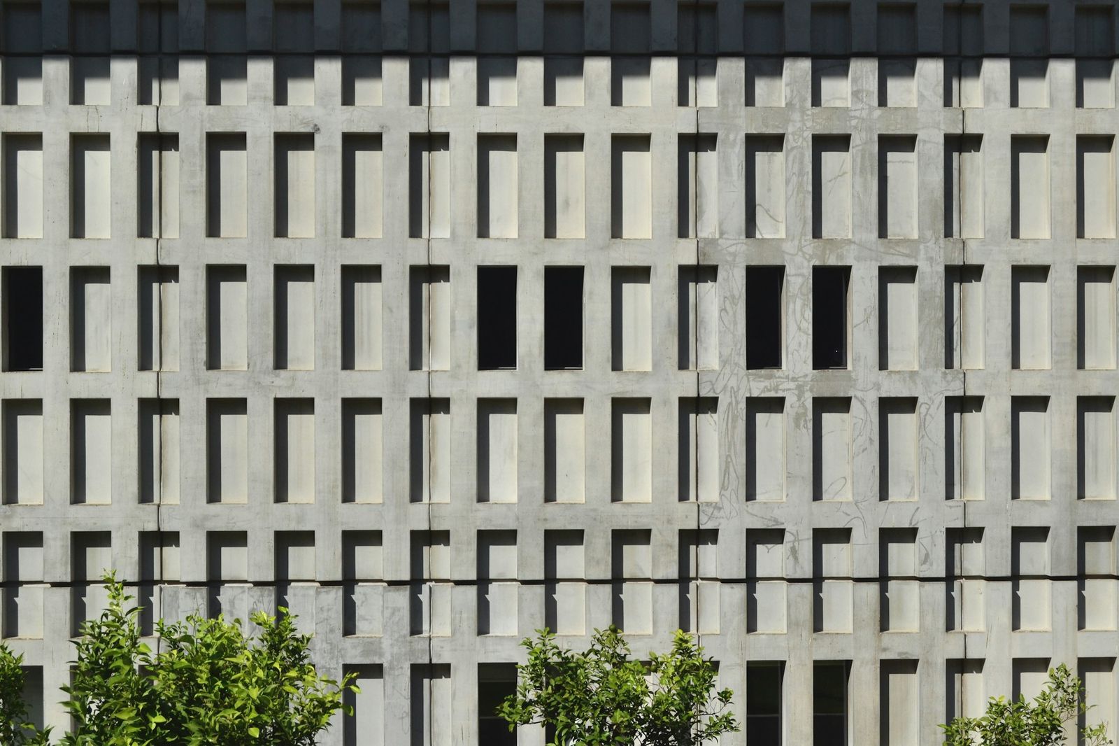 A concrete façade with a deep grid of window openings, small trees along its base.