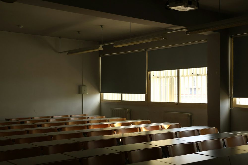 Dim lecture room with rows of wooden desks and blinds half drawn