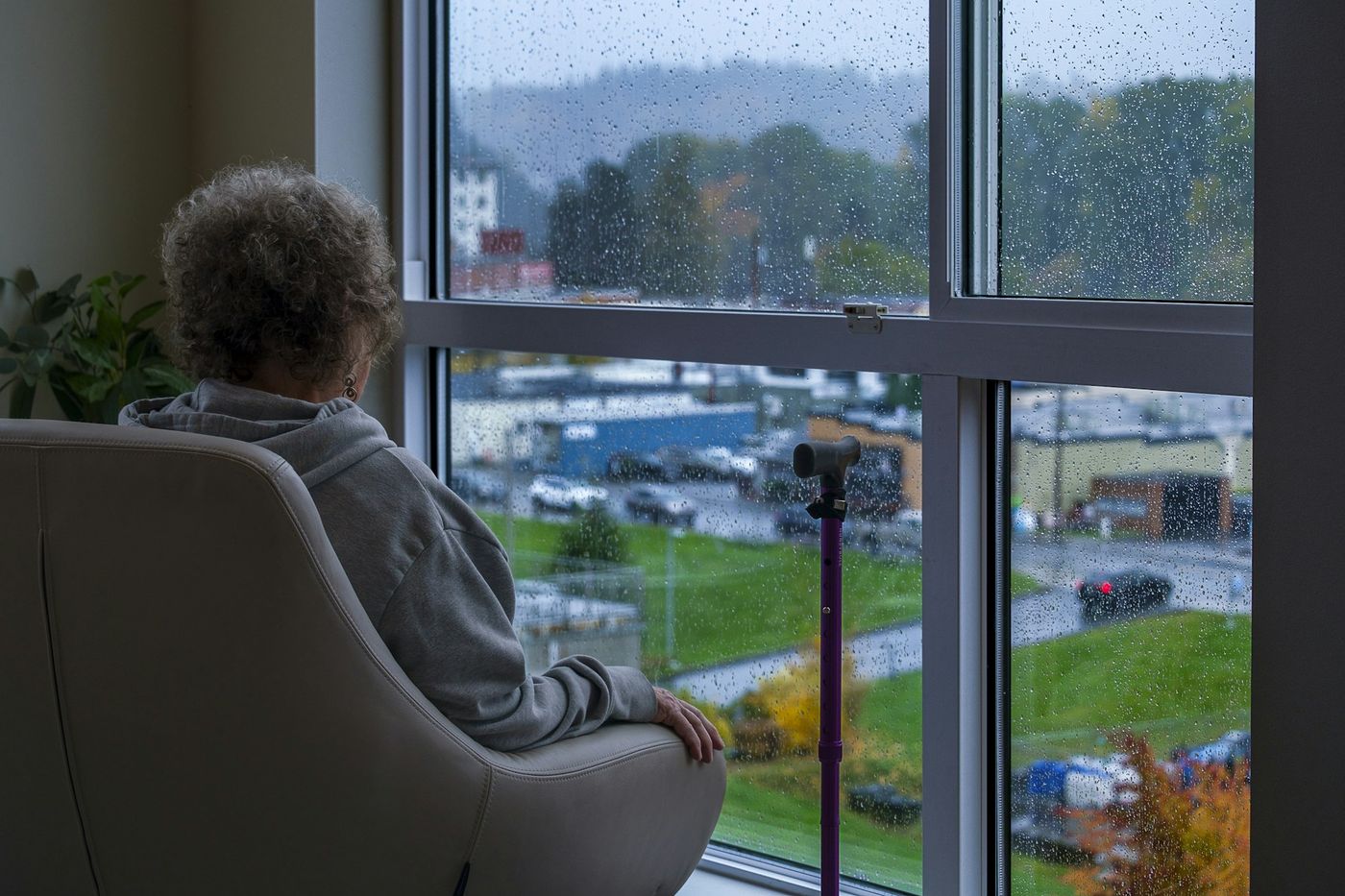 An older person in an armchair watching rain on a large window.