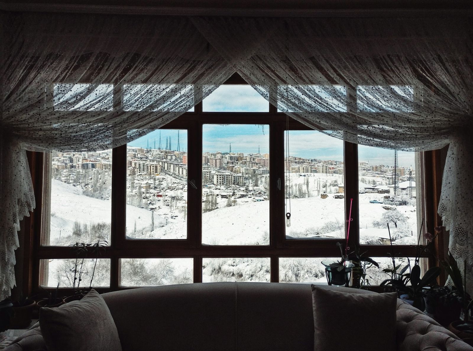 A living room with a sofa facing wide windows onto a snowy hillside town.