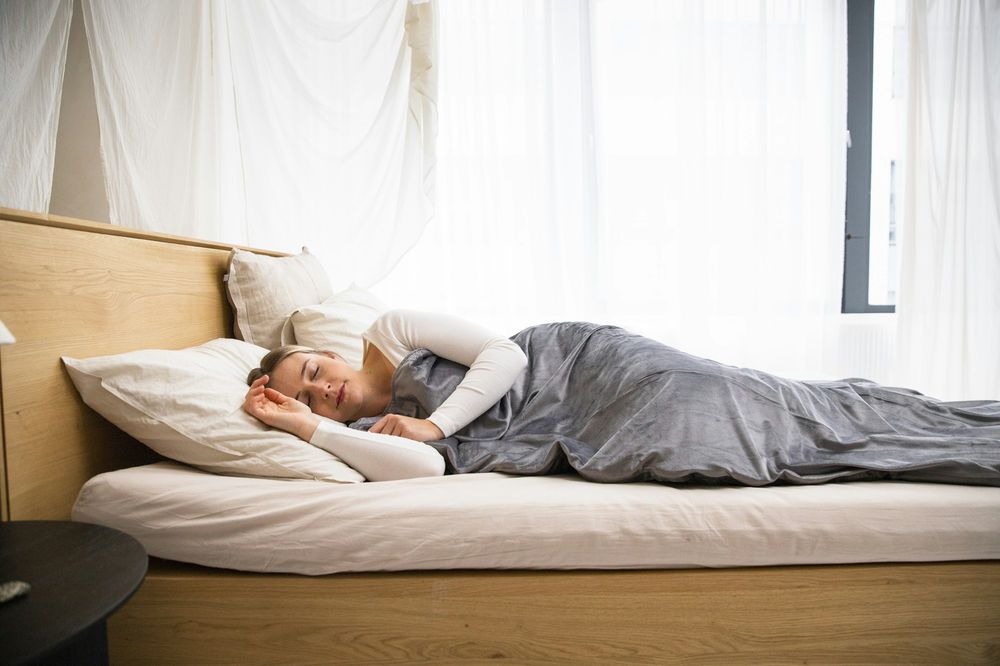 A woman asleep under a grey blanket on a low bed beside a bright window