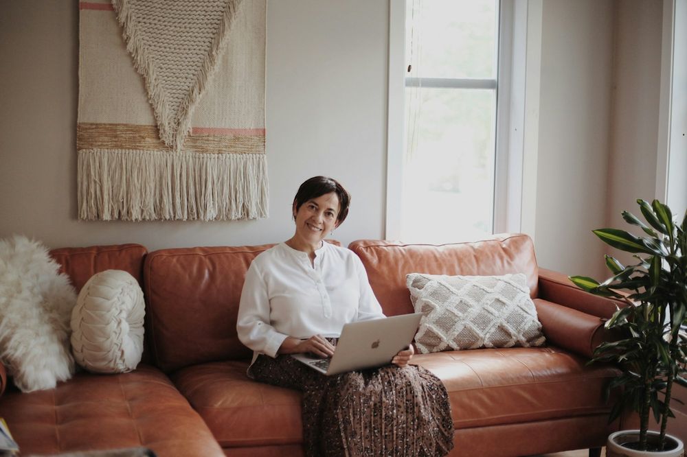 A woman working on a laptop on a tan leather sofa, a woven hanging on the wall behind her