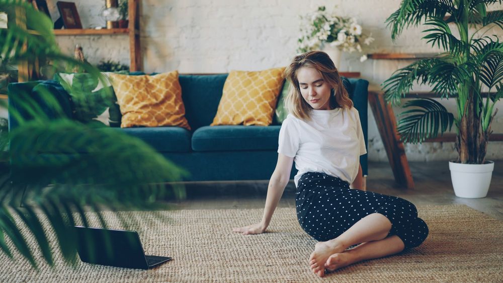 A young woman sitting on a jute rug in front of a teal sofa, with palms around her