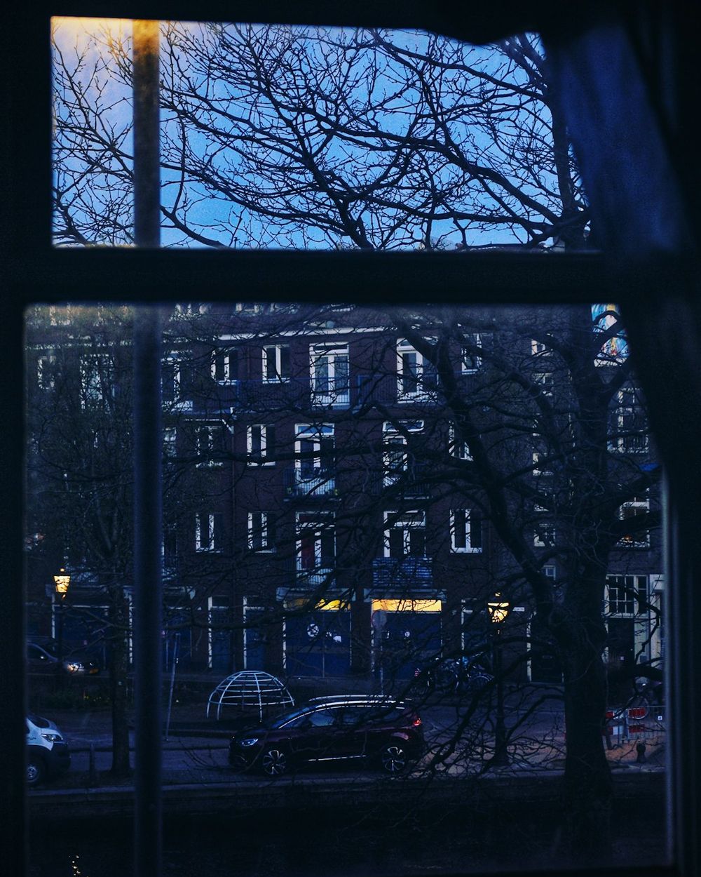 A dark window with bare winter branches outside and a row of houses with a few lit doorways.