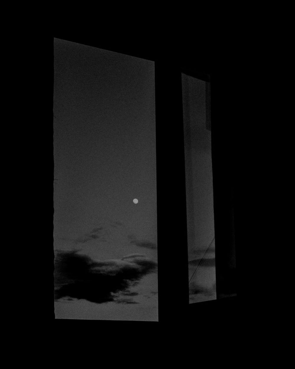 An almost black room with a tall window, a small moon and low clouds in a grey sky.