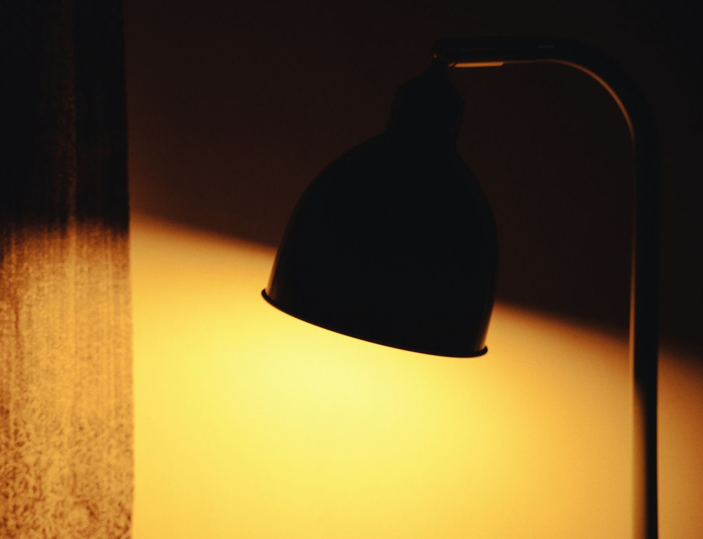 A reading lamp in silhouette, its light falling warm across a dark wall beside a curtain.
