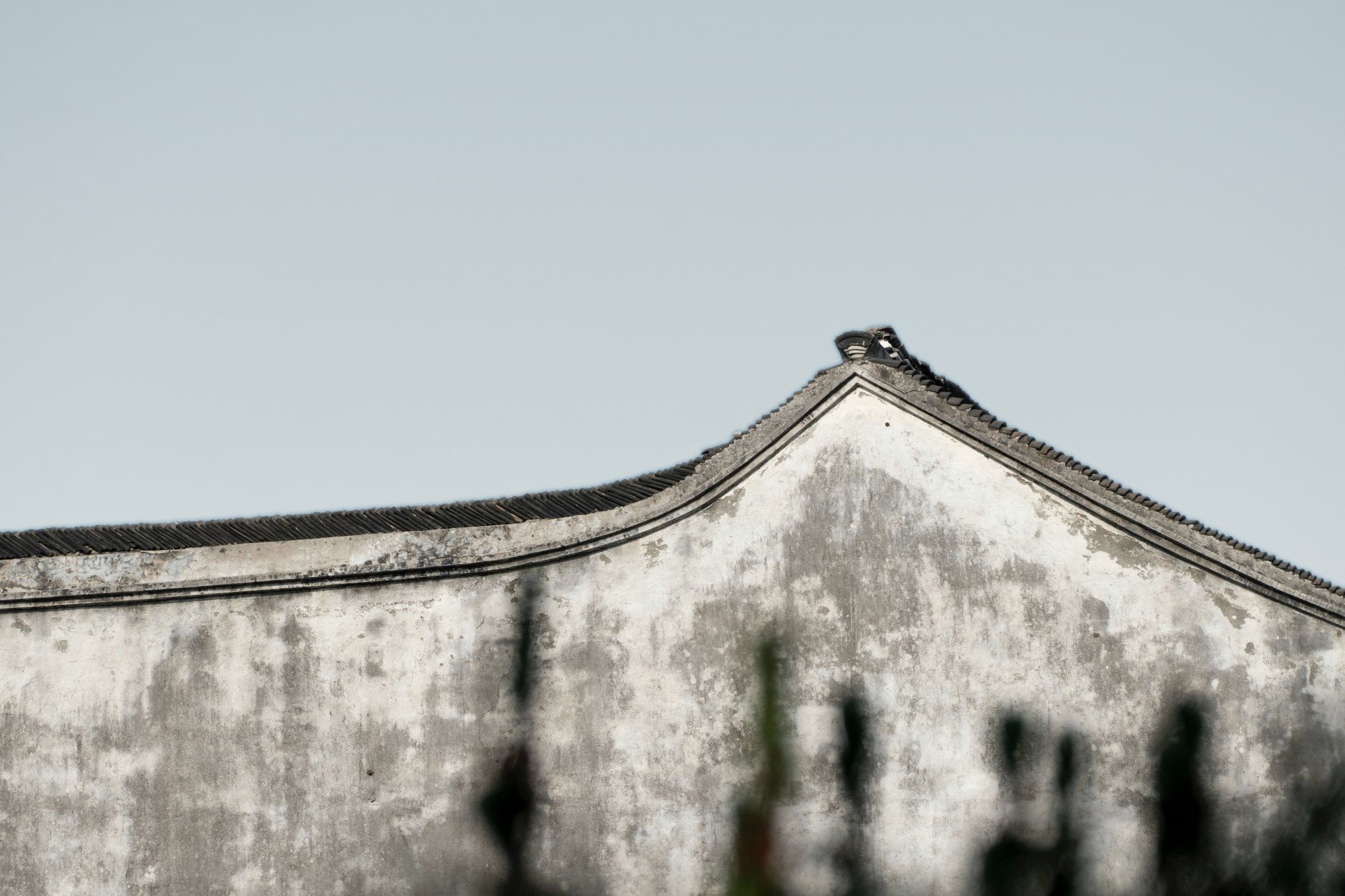 A weathered whitewashed gable wall with a curved grey tile roofline under a pale sky