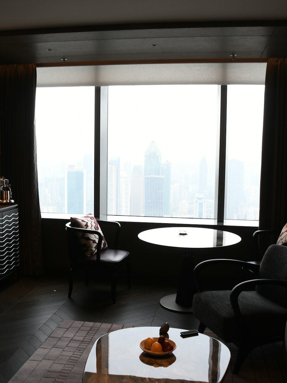 A dark hotel suite high above a city, armchairs and a round table set against a wide window.