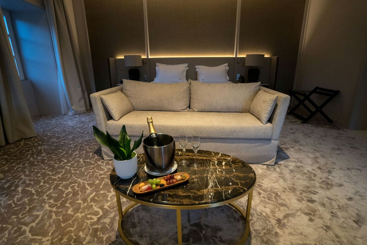 A hotel suite at dusk: a linen sofa, a black marble table with champagne on ice, and lamps glowing behind the bed.