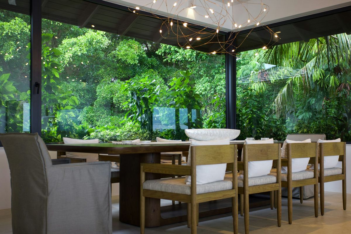 A glass dining pavilion set in a dense garden, a long wooden table under a web of small lights.