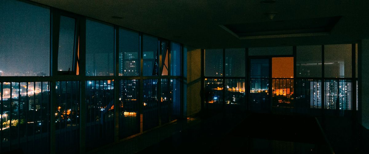 A dark penthouse at night, a wall of windows full of city lights.