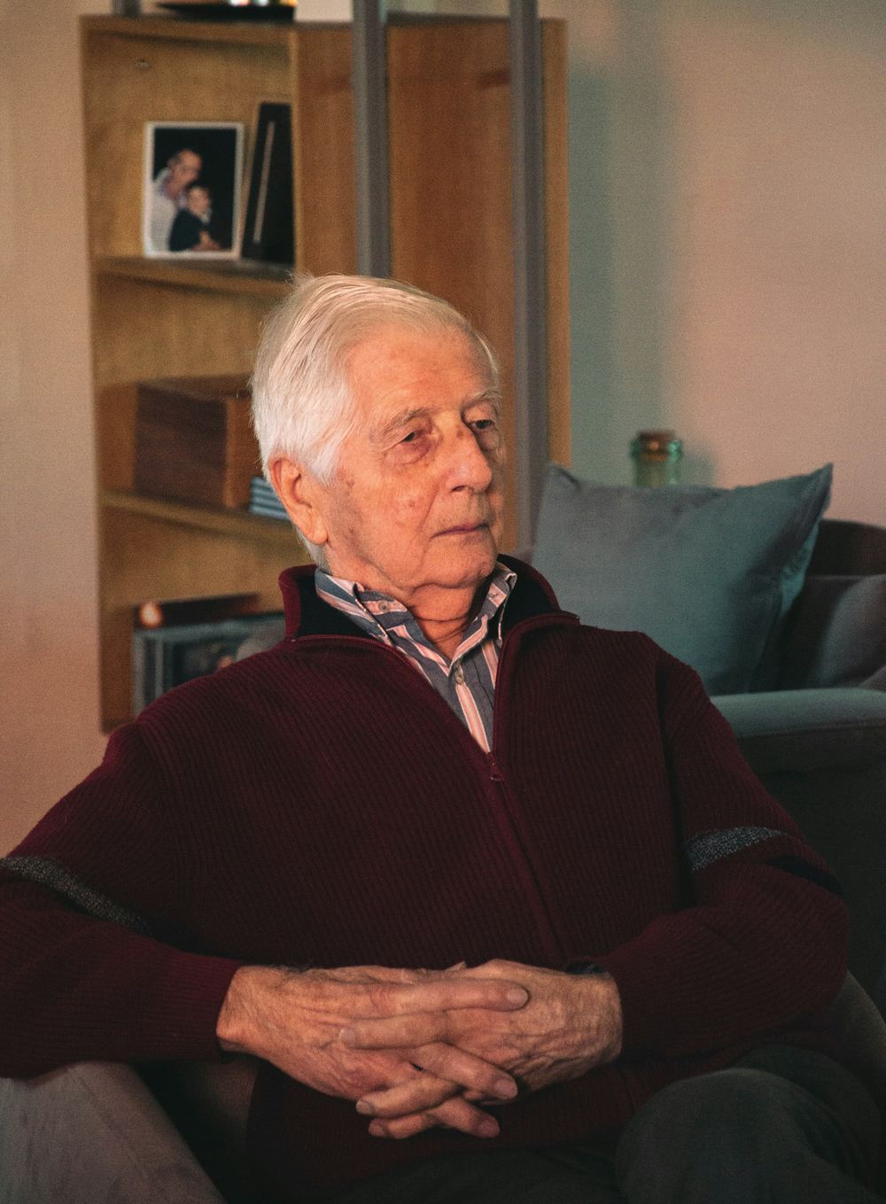 An older man with white hair sits in an armchair at home, hands folded, a bookcase behind him.