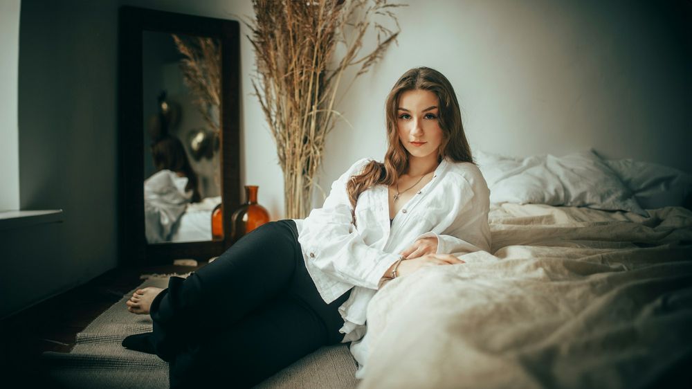 A young woman in a white shirt leans back on an unmade bed in a dim bedroom, dried grasses in a vase behind her.