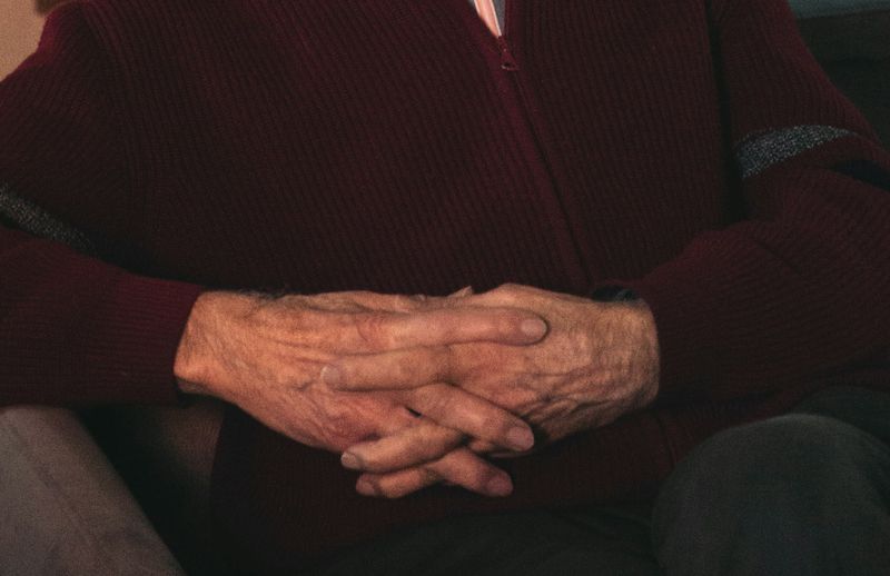 An older man's folded hands resting in his lap, a dark red sweater and a checked shirt cuff.