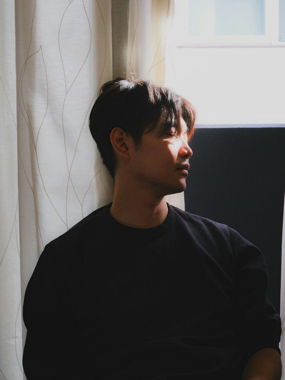 A young man in a dark sweater stands by a window, looking out, curtains behind him.