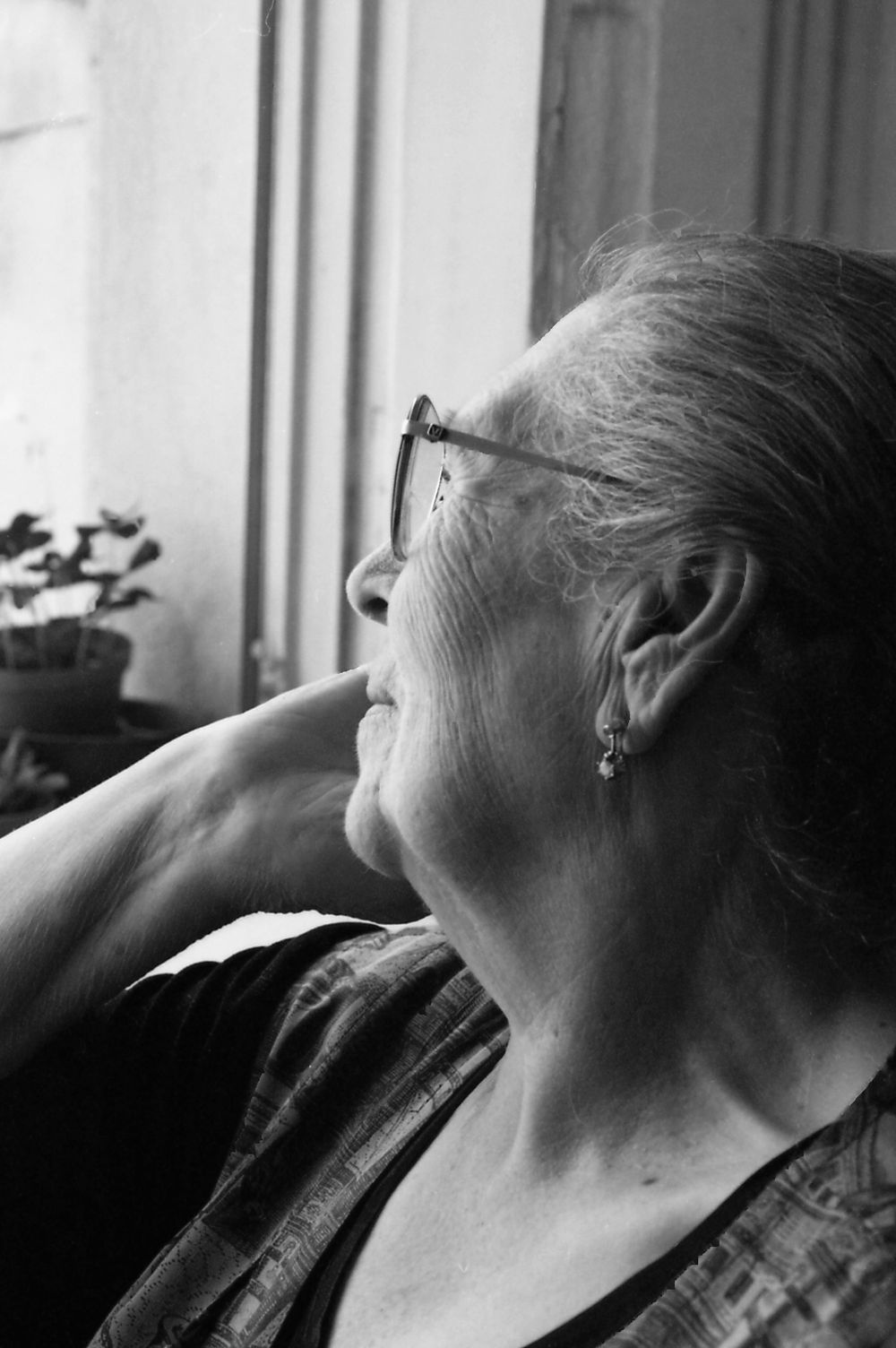 Black and white photograph of an older woman in glasses looking out of a window, her hand at her neck.