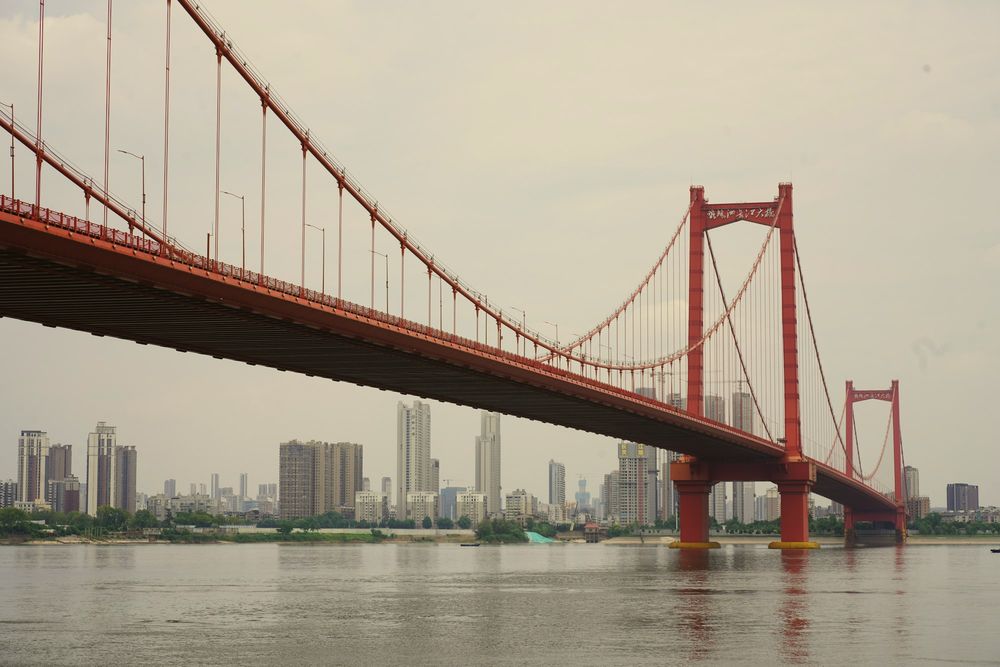 A red suspension bridge over the Yangtze under a grey summer sky, towers on the far bank.