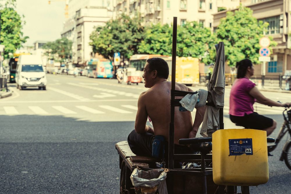 A shirtless man rests on a street cart at a sunny crossing while a woman cycles past.