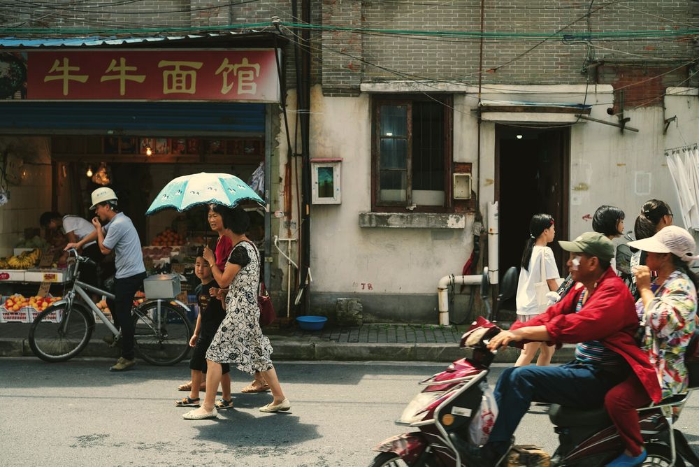 A street outside a noodle shop: a woman walks under a sun umbrella, people pass on scooters.