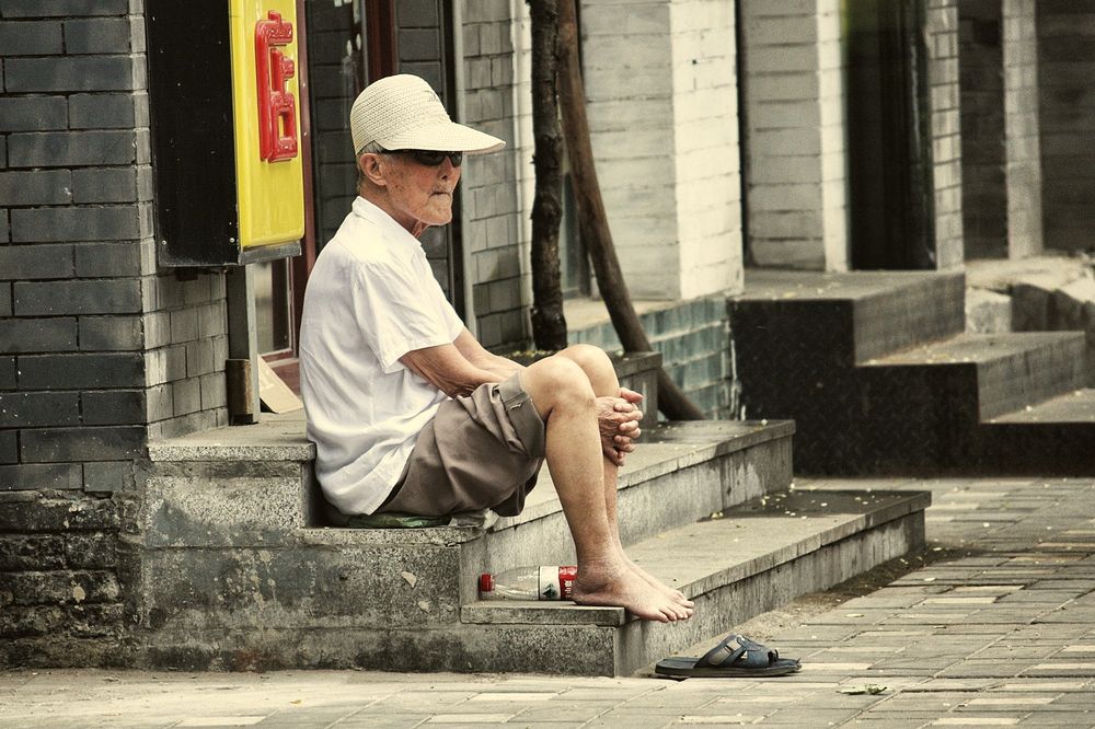 An old man in a white cap sits barefoot on stone steps, his sandals beside him.