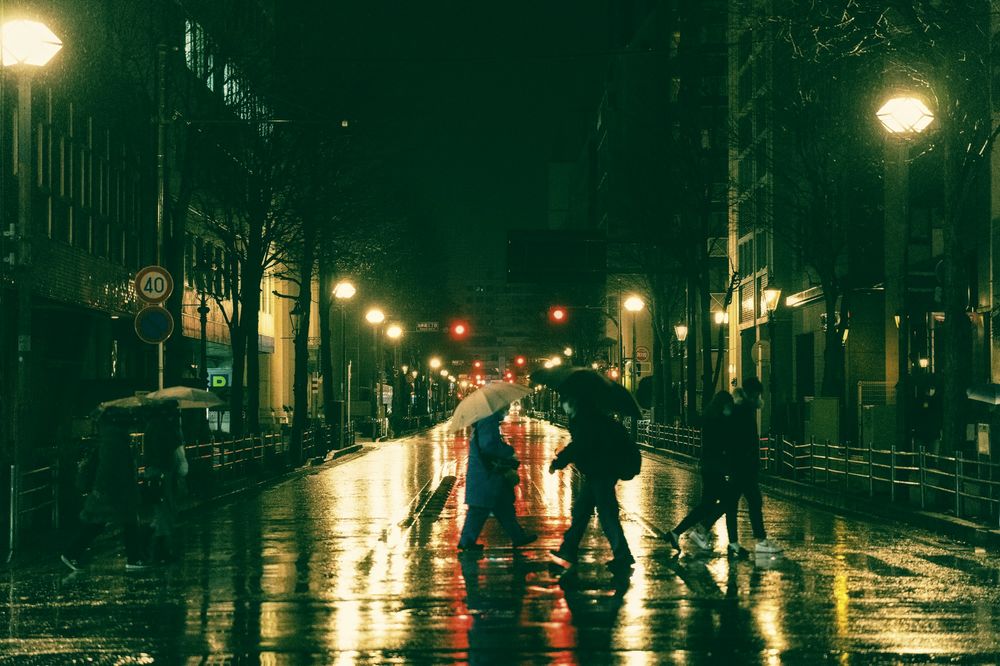 A wet street at night under green-tinted light, people crossing with umbrellas.