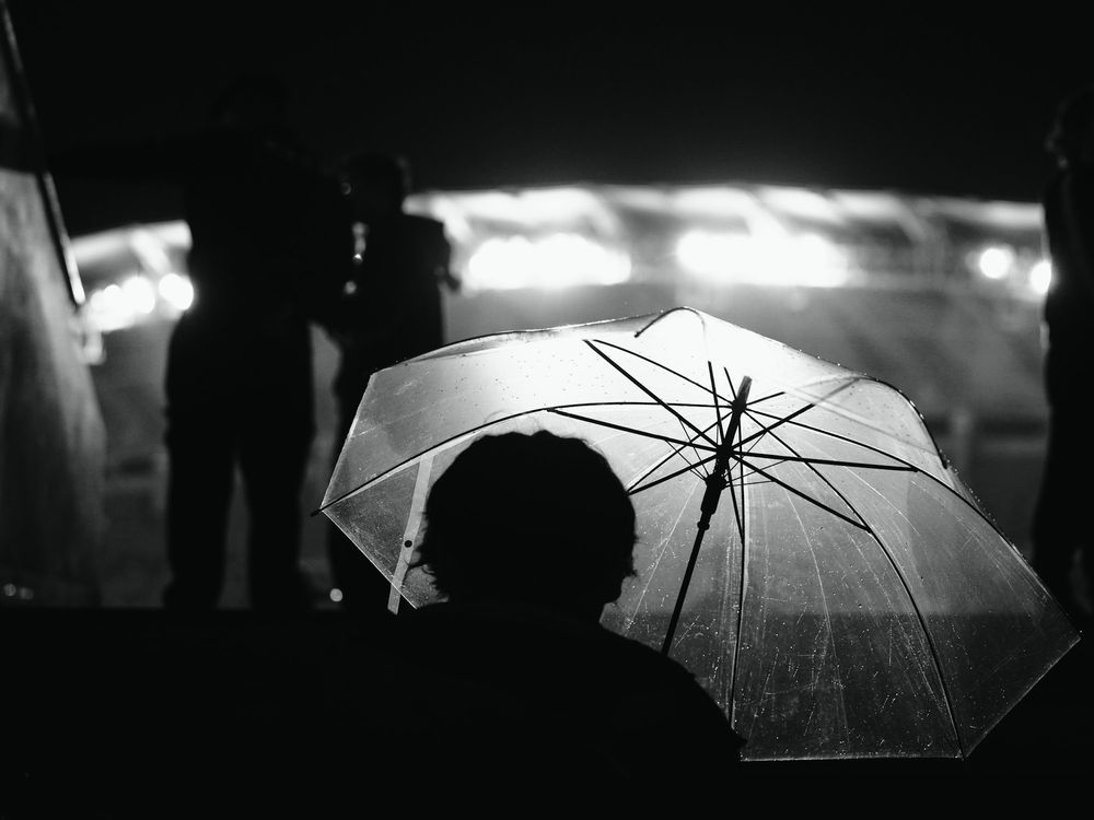 Black and white: a figure under a clear umbrella in the rain, lights blurred behind.