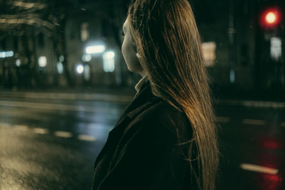 A woman seen from behind on a wet street at night, a red traffic light beyond.