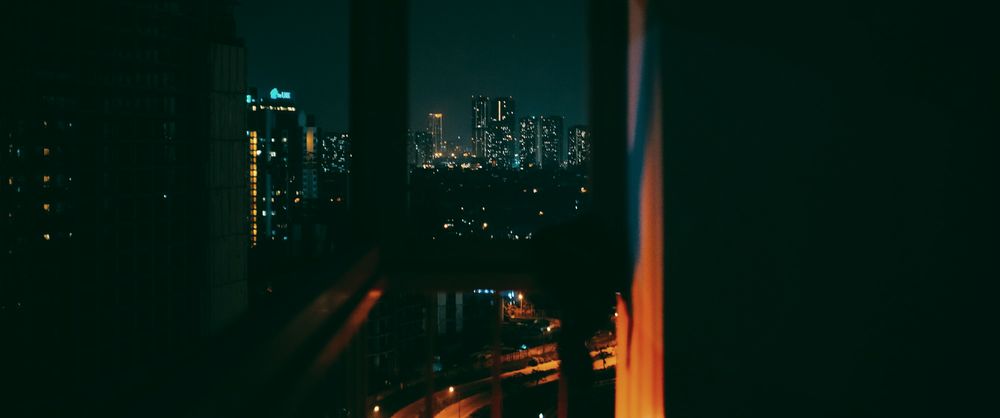 A dark balcony corner at night, a curtain lit orange, towers beyond the window.