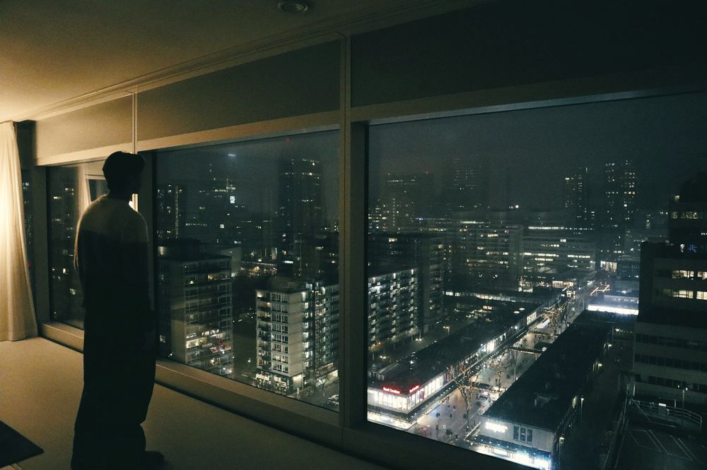A man stands calmly in a dark, quiet room at a tall window above the lit city.