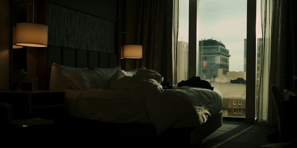 A dim bedroom at dusk: an unmade bed, two lamps, and a tall window onto city buildings.