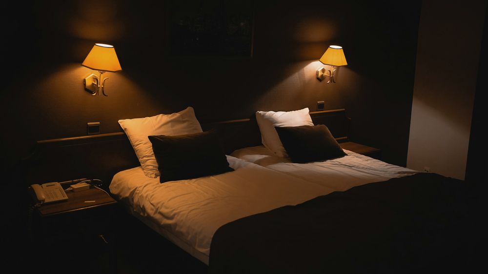 A dark bedroom with white bedding, lit by two warm wall lamps.