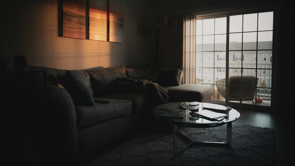 A living room in the evening: a grey sofa, a round glass table and low sun through a balcony door.